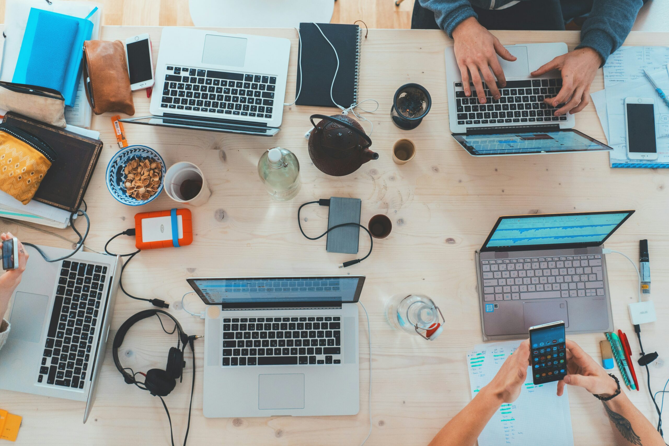 An overhead view of a desk. On the desk, there are computers, hard drives, headphones, and people's hands typing on computers or using phones.